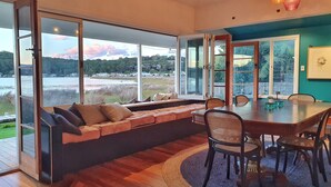 A modern dining area with a large table and chairs, a built-in bench with cushions, and large windows offering a view of a lake and hills. - Reimagined Designer Lakehouse in Prime Location (Burrill Lake)