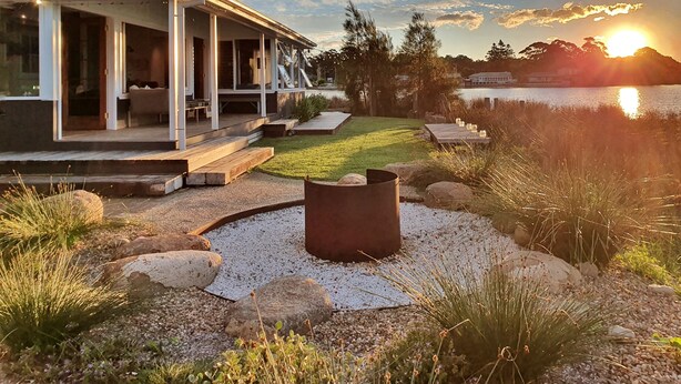 A modern house with a covered porch, a fire pit, and a view of a lake at sunset. - Reimagined Designer Lakehouse in Prime Location (Burrill Lake)