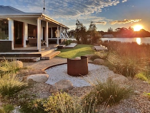 Une maison moderne dotée d’un porche abrité, d’un foyer extérieur et offrant une vue sur un lac au coucher du soleil.