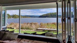 A modern room with a large window overlooking a lake, a cushioned seating area, and a wooden table. - Reimagined Designer Lakehouse in Prime Location (Burrill Lake)