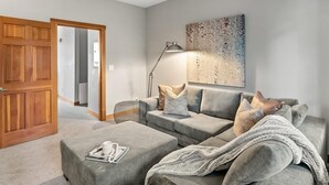 Ein modernes Wohnzimmer mit einem grauen Sofa, einem Couchtisch und einer Lampe. Eine Holztür und ein Gemälde sind an der Wand zu sehen.
