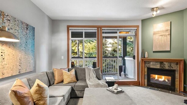 Ein Wohnzimmer mit grauem Sofa, einem Couchtisch und einem Kamin. Ein großes Fenster bietet Blick auf eine grüne Landschaft.