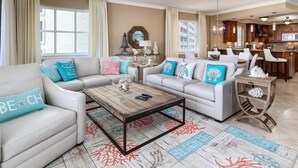 A spacious living room with white sofas, a wooden coffee table, and decorative pillows. A kitchen is visible in the background.