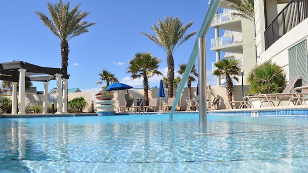 A clear swimming pool with lounge chairs and umbrellas, surrounded by palm trees and a building with balconies.