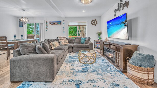 A living room with a sectional sofa, a flat-screen TV, a fireplace, and a large window with a view of the outdoors. - Seaside Riverfront Escape • HotTub • Walk to Beach (Seaside)