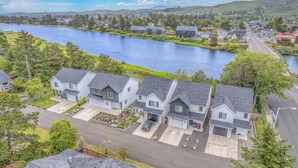 A residential area with houses along a river, featuring a mix of single and two-story buildings, a clear sky, and a hilly landscape in the background. - Seaside Riverfront Escape • HotTub • Walk to Beach (Seaside)