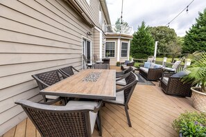 A covered patio with a wooden table and wicker chairs, surrounded by potted plants and a view of the house.