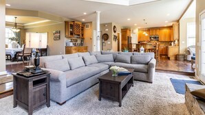 A spacious living room with a sectional sofa, a coffee table, and a dark wooden side table. A kitchen with wooden cabinets and a dining area with a table and chairs are visible in the background.