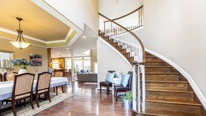 A spacious living area with a wooden staircase, a dining area with a table and chairs, and a kitchen in the background.