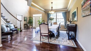 A spacious living room with hardwood flooring, a dining area with a table and chairs, a staircase, and a large window with curtains.