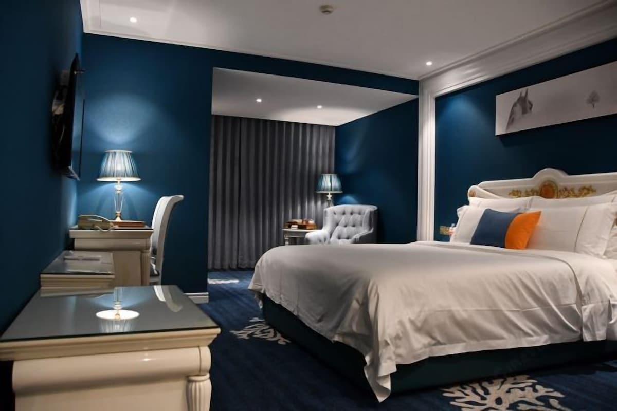 A bedroom with a large bed, a desk, two lamps, a chair, and a curtain.