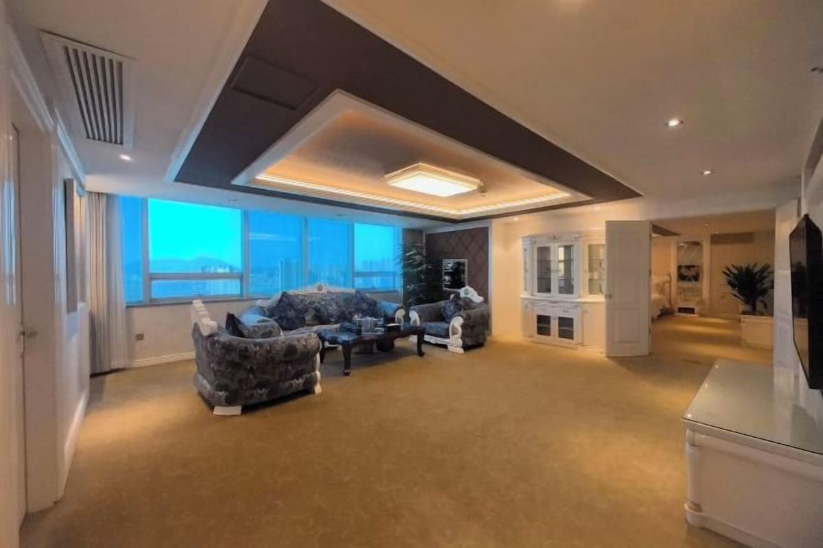 A spacious living room with a high ceiling, large windows, and a fireplace.