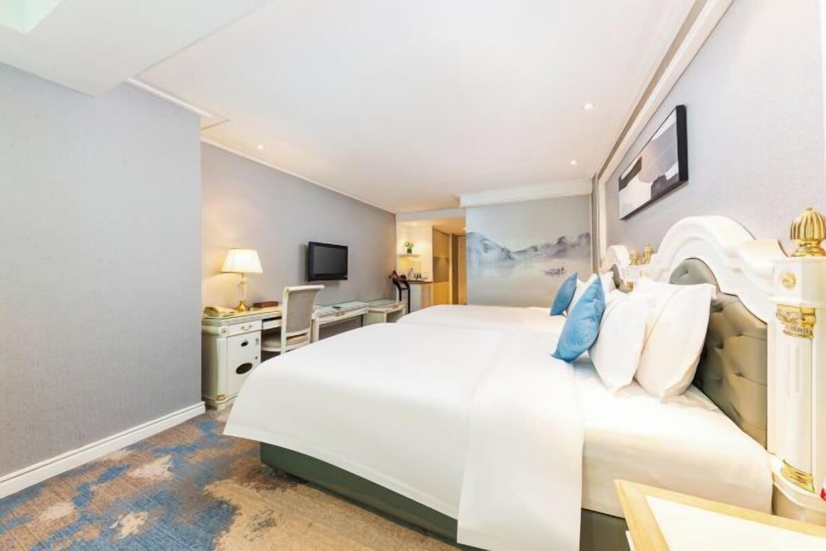 A hotel room with a large bed, a desk with a chair, a television, and a painting on the wall.