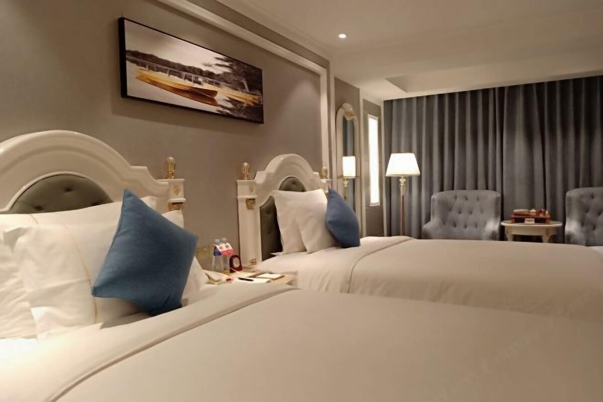 Une chambre d’hôtel avec deux lits, une grande fenêtre avec des rideaux, un tableau au mur et une petite table avec une lampe.