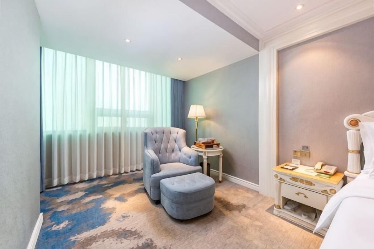 A hotel room with a large window, a grey armchair, a small ottoman, a bedside table with a lamp, and a bed with a white headboard.