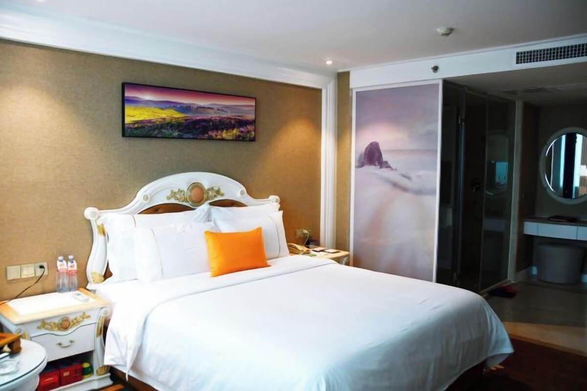 A hotel room with a large bed, a nightstand, a bedside table, a mirror, and a view of a mountainous landscape through a glass door.
