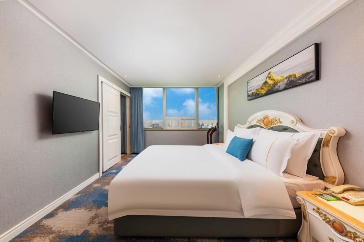 A hotel room with a large bed, a flat-screen TV, a window with a city view, and a painting on the wall.