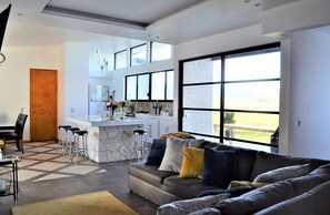 A modern living room with a sofa, a dining area with a table and chairs, a kitchen with a bar, and large windows offering a view. - Casa Lynda's Valle de Guadalupe (Villa de Juárez)
