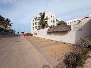 A street view with a building featuring a thatched roof, palm trees, and a sign indicating a rental property.