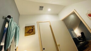 Une pièce avec une porte blanche, un tableau représentant un oiseau, une bouche d’aération au plafond, et un aperçu d’un coin repas avec une table et des chaises.