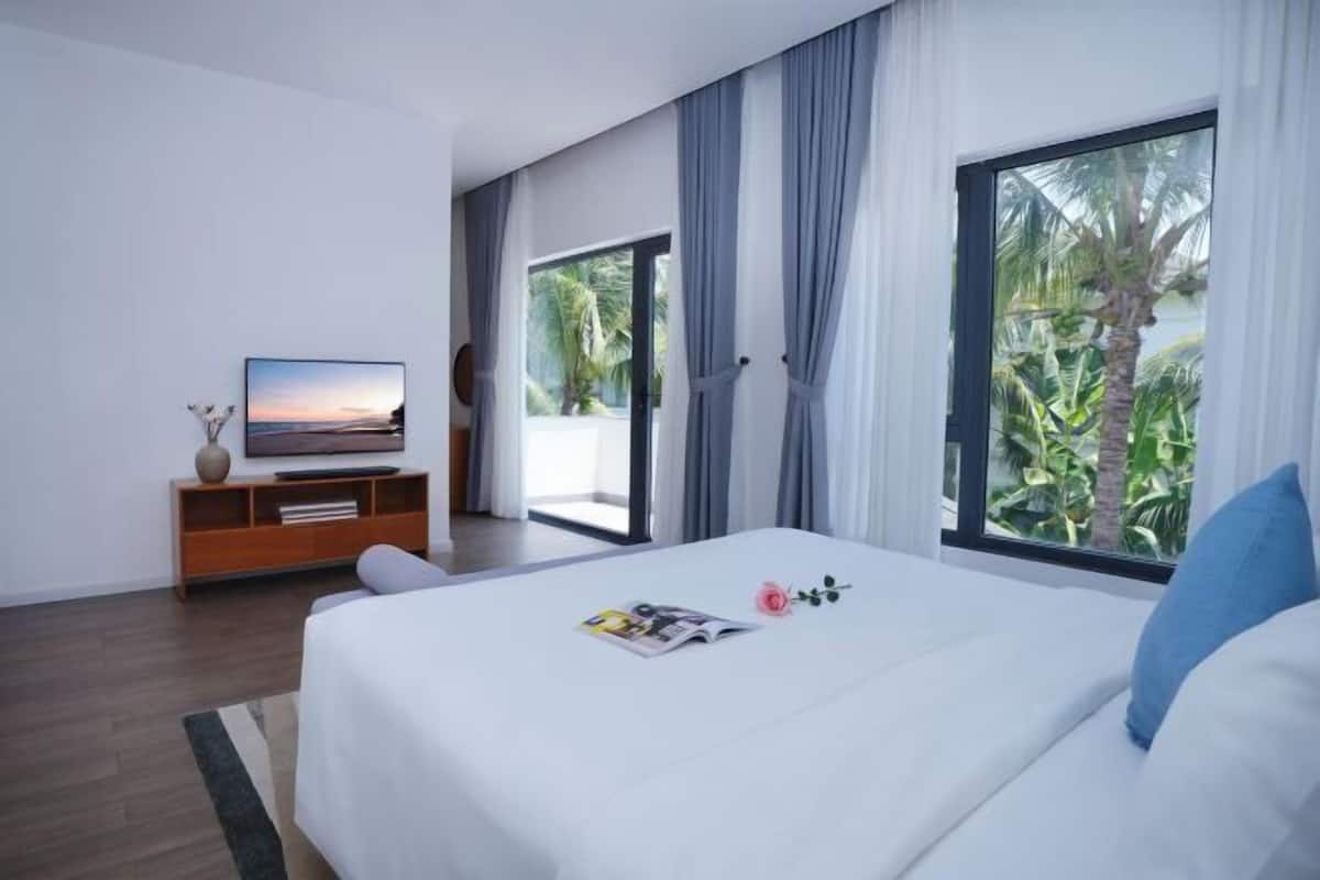 A modern hotel room with a large bed, a television on a wooden stand, and a view of palm trees through the window.