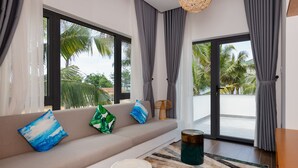 A modern living room with a sofa, pillows, a round table, and a view of palm trees through large windows. - Le Palmier Ho Tram Resort (Ho Chi Minh City)