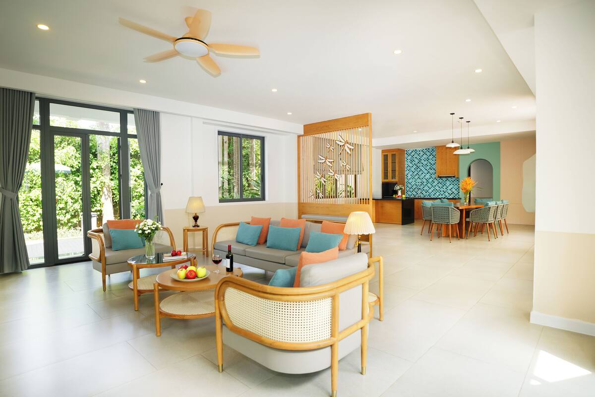 A spacious living area with a sofa, coffee table, and dining table, connected to a kitchen with a view of greenery.
