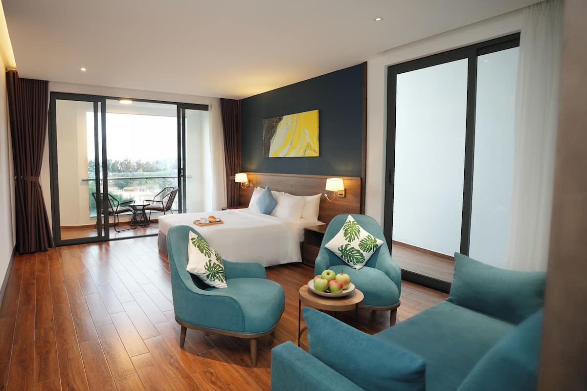 A hotel room with a bed, two armchairs, a small table with fruit, a balcony with a view, and a painting on the wall.