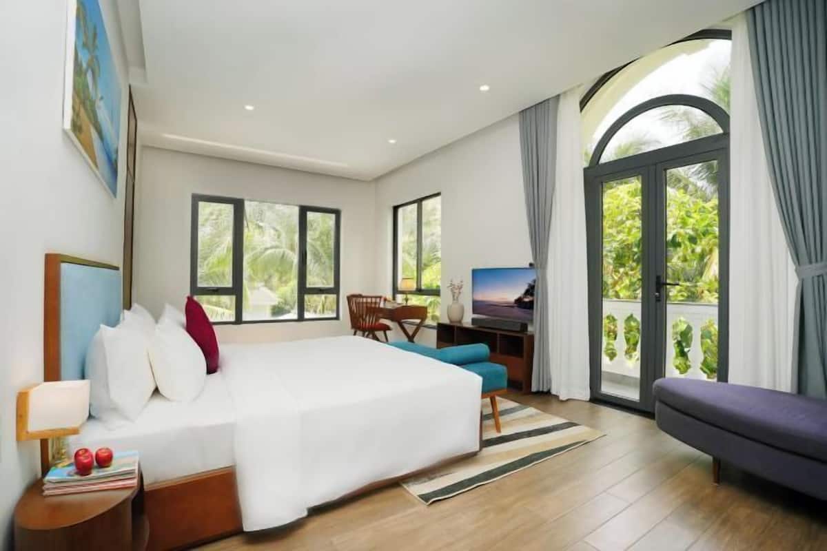 A modern hotel room with a large bed, a desk, and a balcony with a view of greenery.