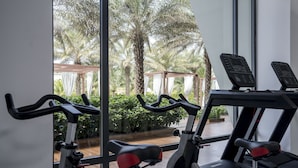 A gym with stationary bikes and treadmills, large windows with a view of palm trees and a wooden deck.