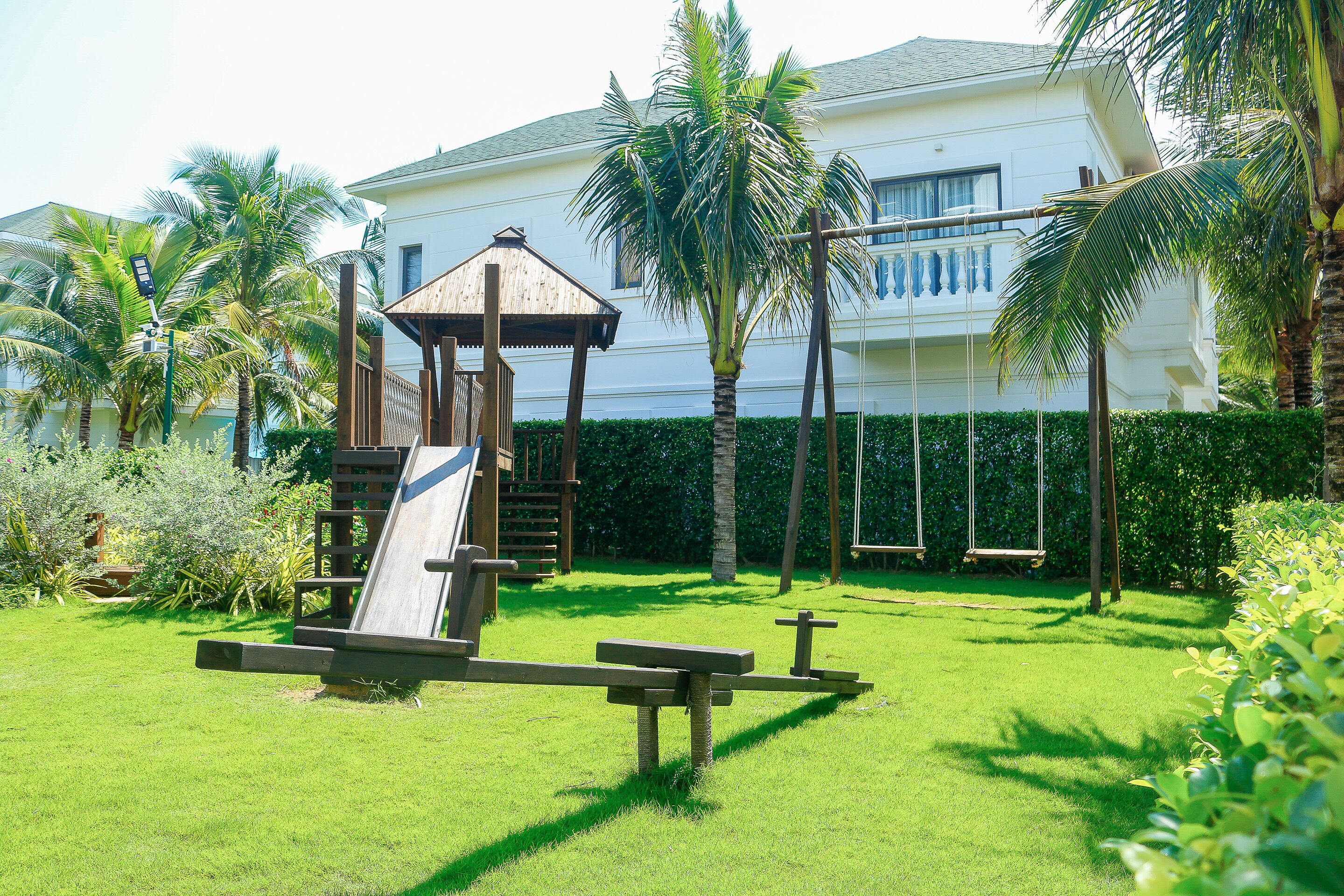 A playground with a slide and swings, surrounded by a well-maintained lawn and palm trees, with a building in the background.