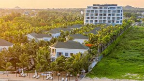 An aerial view of a beachfront hotel with a large building, multiple villas, and a beach with lounge chairs. - Le Palmier Ho Tram Resort (Ho Chi Minh City)