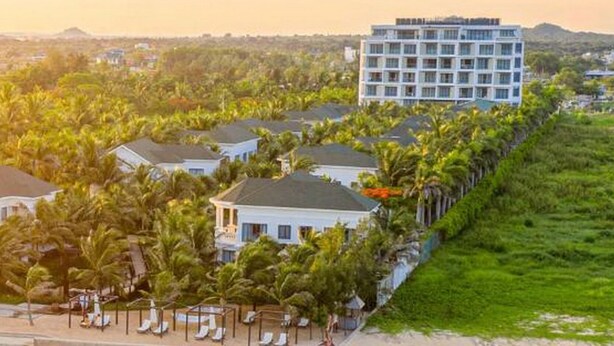 An aerial view of a beachfront hotel with a large building, multiple villas, and a beach with lounge chairs.
