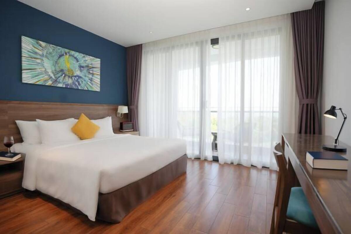 A hotel room with a bed, a desk, a chair, a lamp, a painting on the wall, and a window with curtains.