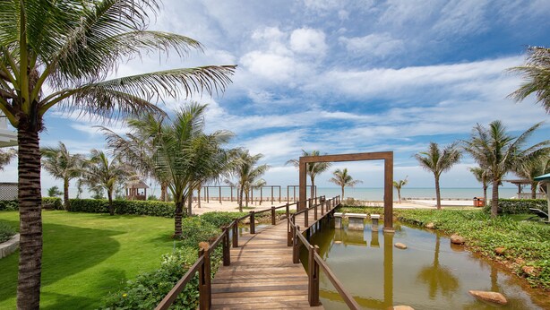 A wooden bridge over a water feature with palm trees and a beach in the background. - Le Palmier Ho Tram Resort (Ho Chi Minh City)