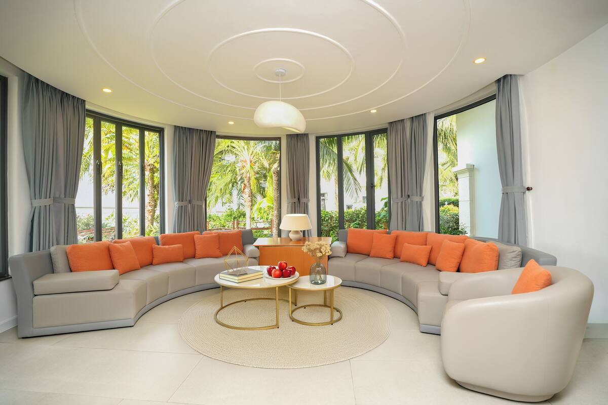 A modern living room with a curved sofa, two side tables, a lamp, and a view of greenery through large windows.