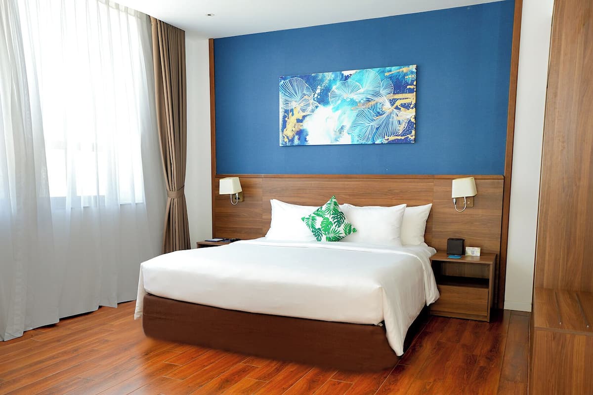 A hotel room with a large bed, wooden headboard, two bedside tables with lamps, a blue accent wall, and a window with sheer curtains.