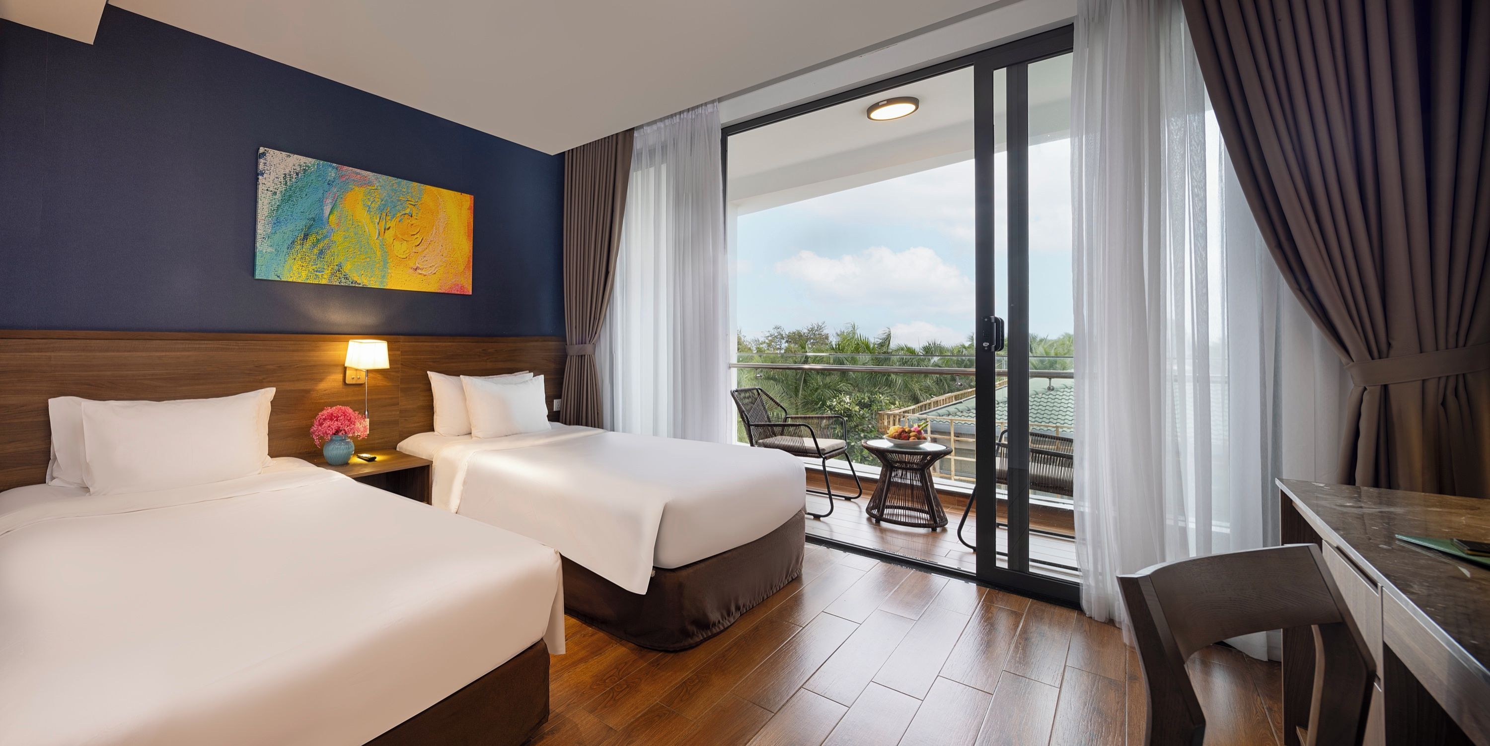 A hotel room with two beds, a nightstand with a lamp, a painting on the wall, and a balcony with a table and chairs.