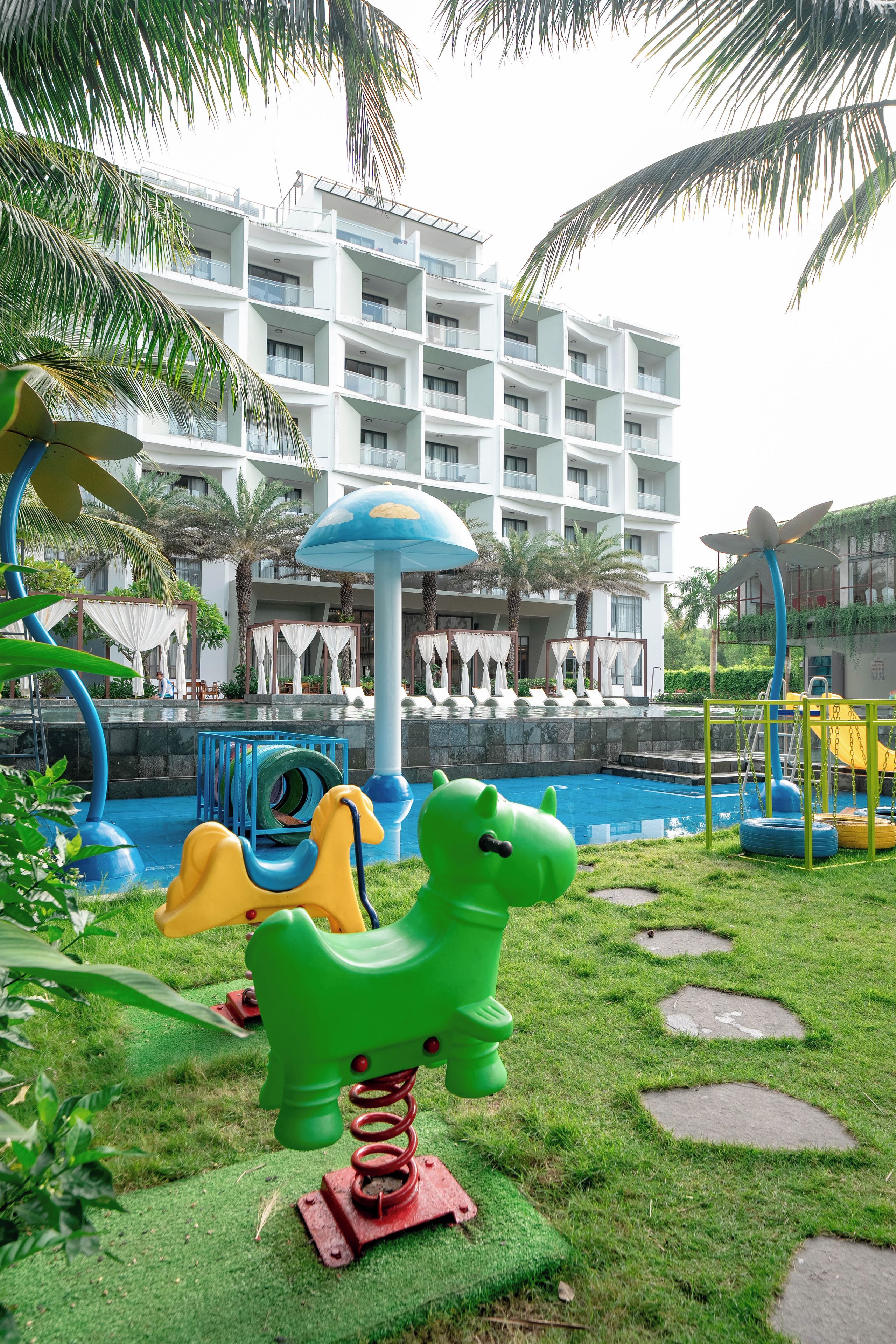 Zona de juegos infantiles con juguetes inflables y piscina, rodeada de palmeras y un edificio de hotel.