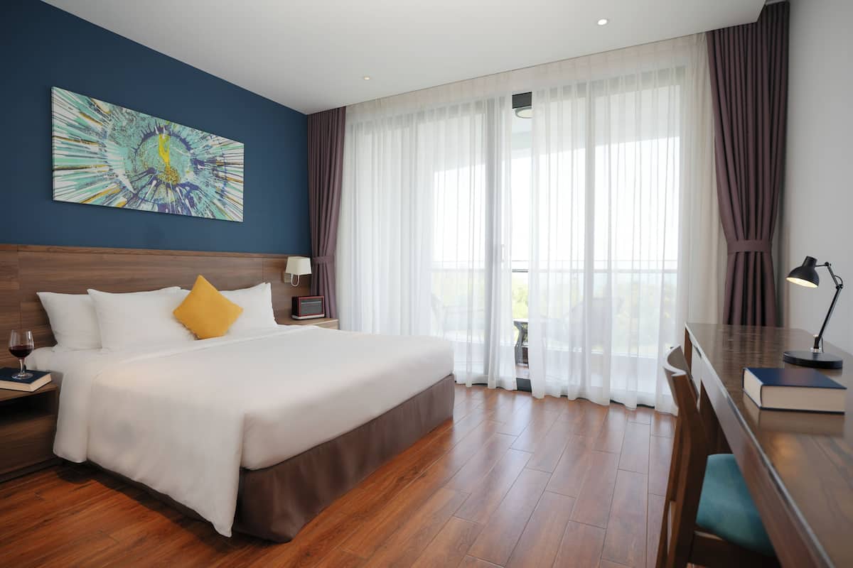 A hotel room with a bed, a desk, a chair, a lamp, a painting on the wall, and a view through the window.