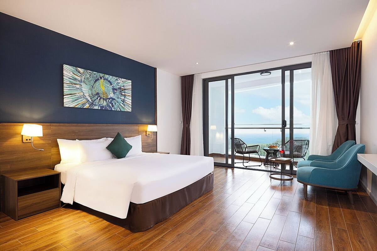 A hotel room with a large bed, a view of the ocean, a balcony with seating, and a painting on the wall.