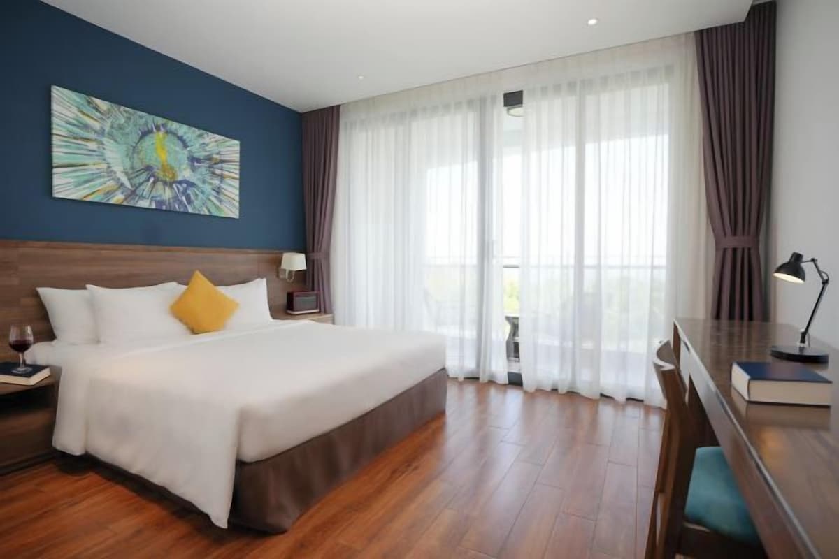 A hotel room with a large bed, a desk, and a view of the outdoors through sheer curtains.