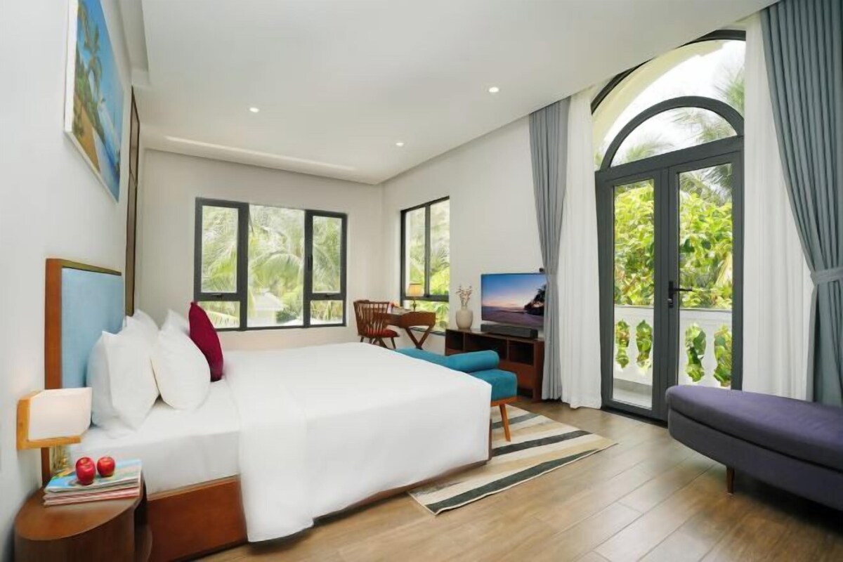 A modern hotel room with a large bed, a desk, and a balcony with a view of greenery.