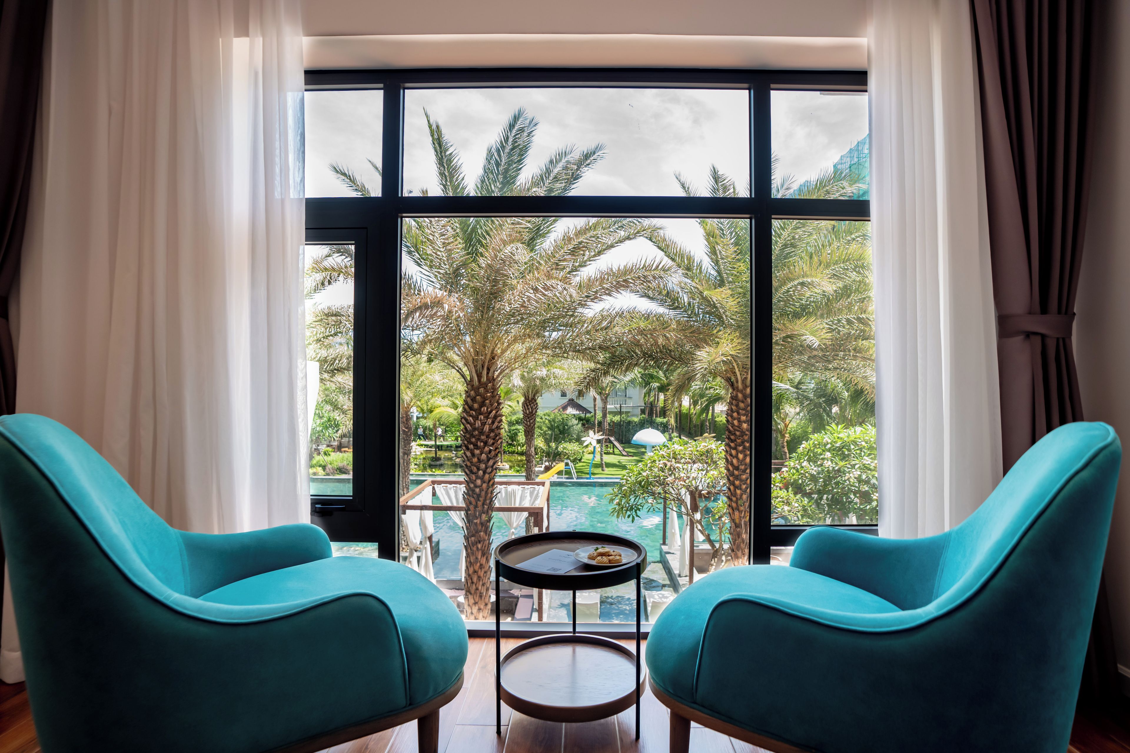 Una habitación con dos sillas color turquesa, una pequeña mesa redonda y un ventanal con vistas a una piscina y palmeras.