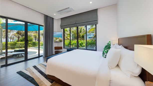 A modern hotel room with a large bed, a view of a pool and palm trees, and a sliding glass door to the outdoor area. - Le Palmier Ho Tram Resort (Ho Chi Minh City)