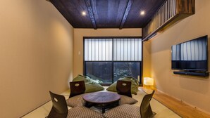 A room with tatami mat seating, a round table, a TV mounted on the wall, and a large window with a view of trees.