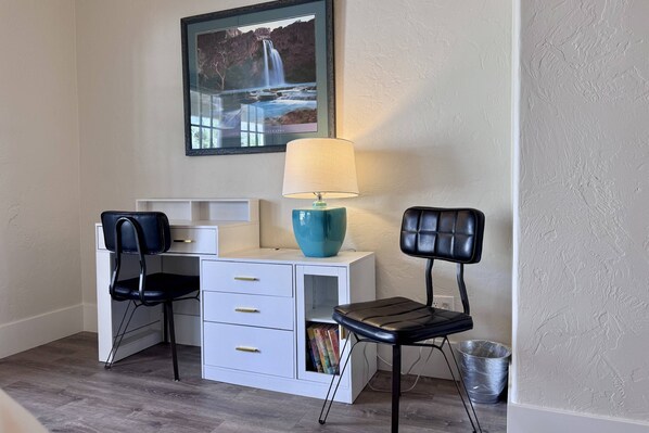 A room with a desk, two chairs, a lamp, a framed picture, and a trash can.