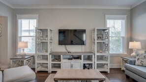 Ein Wohnzimmer mit an der Wand montiertem Fernseher, einem Holztisch und weißen Regaleinheiten mit Dekoration.