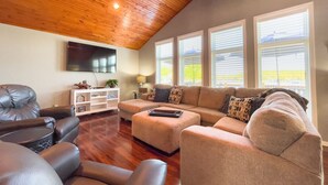 Un salon doté d’un plafond en bois, d’un grand téléviseur à écran plat, d’un ensemble de canapés beige et d’un meuble de rangement blanc pour les appareils multimédias.