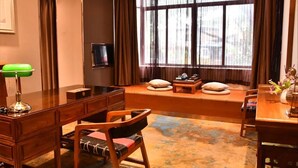 Ein gemütliches Hotelzimmer mit Schreibtisch aus Holz, einem bequemen Sessel, einem Fernseher und einem großen Fenster mit Vorhängen.